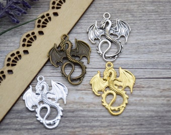 May include: Four metal dragon charms in silver, bronze, and gold tones. Each charm features a detailed dragon design with outstretched wings and a loop for attaching to jewelry or other crafts. The charms are displayed on a wooden surface.