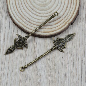 K&ouml;nnte beinhalten: Zwei bronzefarbene Speeranh&auml;nger mit aufwendigen Designs. Jeder Anh&auml;nger hat einen detaillierten Speerkopf und einen strukturierten Schaft, ideal f&uuml;r Schmuckherstellung oder Bastelprojekte. Die Anh&auml;nger liegen auf einem Holzuntergrund.