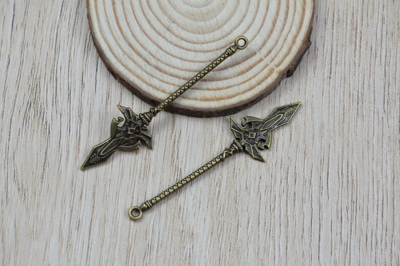 K&ouml;nnte beinhalten: Zwei bronzefarbene Speeranh&auml;nger mit aufwendigen Designs. Jeder Anh&auml;nger hat einen detaillierten Speerkopf und einen strukturierten Schaft, ideal f&uuml;r Schmuckherstellung oder Bastelprojekte. Die Anh&auml;nger liegen auf einem Holzuntergrund.