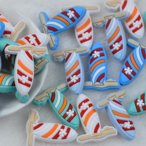 Op de afbeelding: Een verzameling kleurrijke bedels in de vorm van een surfboard. De bedels hebben verschillende kleuren, waaronder wit, rood, oranje, blauw en teal. Elke bedel heeft een klein wit bloemdesign en een bruin gekleurde vin.