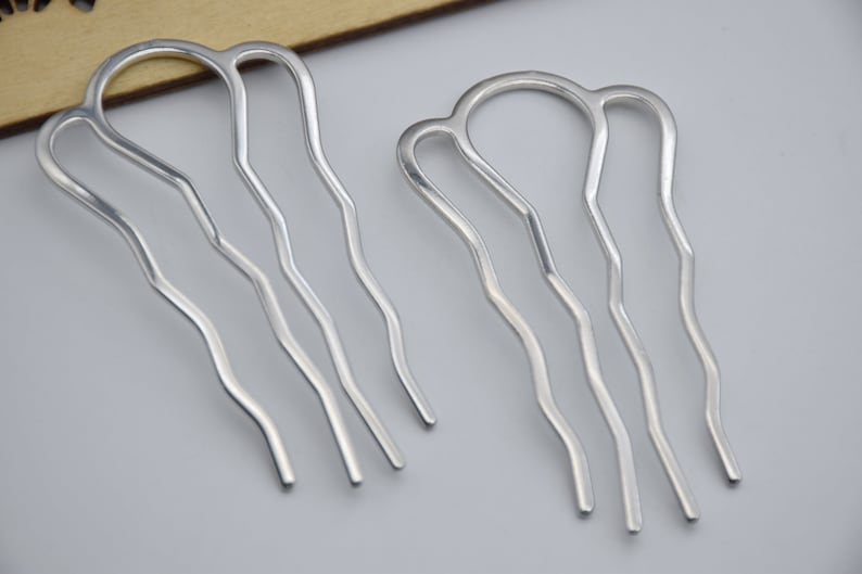 Op de afbeelding: Twee zilveren haar kammen met golvende, decoratieve ontwerpen. De kammen zijn gemaakt van metaal en hebben een glanzende afwerking.