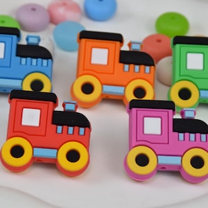 Puede incluir: Una colección de coloridos dijes de tren de juguete en azul, naranja, verde, rojo y rosa. Cada tren tiene un techo negro, una ventana cuadrada blanca y ruedas amarillas. Otras cuentas de colores están esparcidas alrededor de los trenes.