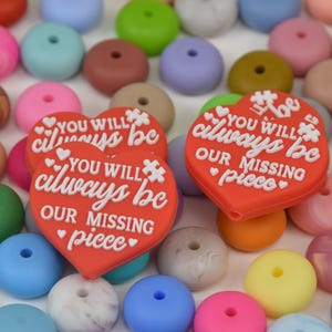 May include: A collection of colorful silicone beads in various shades, including pink, blue, green, and yellow. Two red heart-shaped beads with the text "YOU WILL always be OUR MISSING piece" are featured prominently.