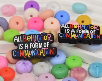 May include: A collection of colorful silicone beads in various shades of pink, purple, blue, green, and yellow. Two black rectangular charms with the text "ALL BEHAVIOR IS A FORM OF COMMUNICATION" in colorful letters are also present.