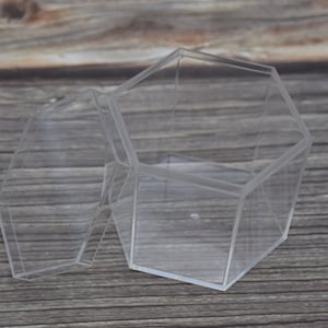 Puede incluir: Una caja hexagonal de plástico transparente con tapa. La caja está abierta y la tapa está apoyada junto a ella. La caja está sobre una superficie de madera.