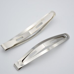 May include: Two silver metal hair clips. The clips are about 5 inches long and have a slight curve to them. They have a single hole in the center of each clip.