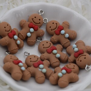 May include: A collection of nine gingerbread man charms with red bows, white buttons, and blue sprinkles. The charms are made of brown polymer clay and have silver jump rings.