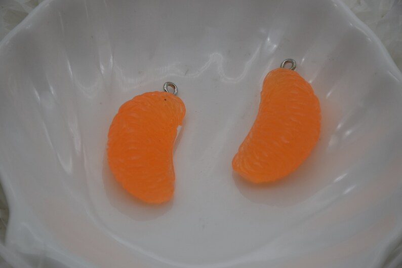 May include: Two orange, tangerine-shaped charms with silver-colored loops for jewelry making. The charms are bright orange and have a textured surface, resembling a real tangerine segment. They are set against a white background.