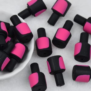 Könnte beinhalten: Eine Sammlung von Miniatur-Nagellackflaschen-Anhängern. Jeder Anhänger hat eine schwarze Flasche mit einem rosa Nagellack-Design. Die Anhänger sind auf einer weißen Oberfläche verstreut, einige in einer kleinen weißen Schale.