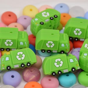 May include: A collection of green silicone baby teethers shaped like recycling trucks. The trucks have a white recycling symbol on the side. The teethers are surrounded by colourful silicone beads.