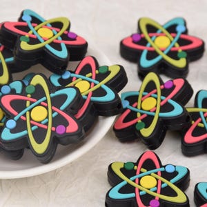 May include: A selection of black atom-shaped charms with vibrant colours. Each charm has a yellow centre, encircled by pink, teal, and lime green orbital rings, and small blue and green dots. The charms are displayed on a white surface.