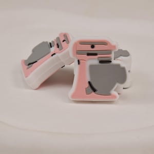 May include: Two decorative charms in the shape of a pink and gray stand mixer and a pink and gray sneaker. The charms are white with pink and gray accents. The charms are likely for shoes or other accessories.