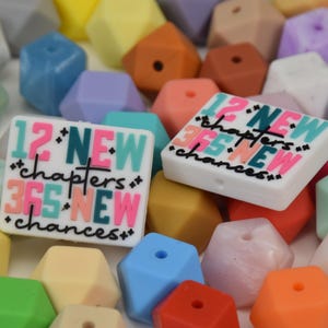 May include: A collection of colorful, geometric beads in various pastel shades, including pink, blue, yellow, and orange. Two white rectangular beads feature the text "12 NEW chapters 365 NEW chances" in a mix of colors and fonts.