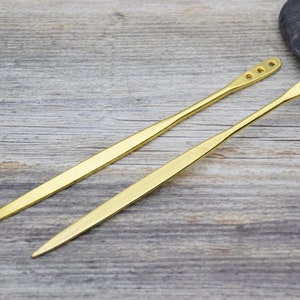 Op de afbeelding: Twee gouden metalen haarstokken met afgeronde uiteinden en meerdere gaten. De stokken liggen op een houten oppervlak.