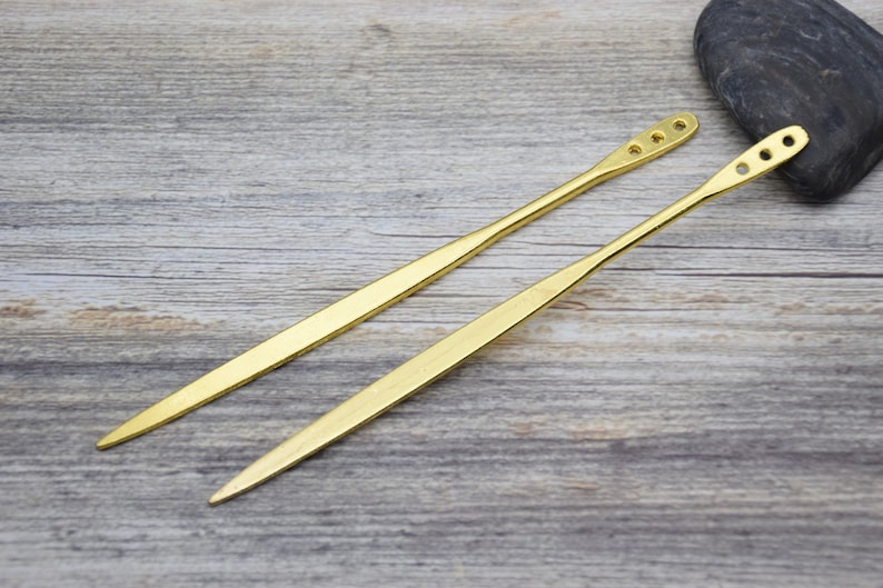 Op de afbeelding: Twee gouden metalen haarstokken met afgeronde uiteinden en meerdere gaten. De stokken liggen op een houten oppervlak.