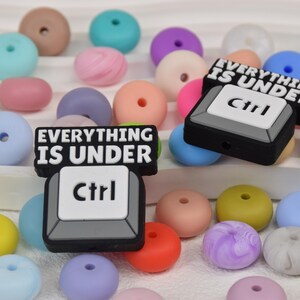 Puede incluir: Una colección de cuentas de silicona coloridas en varios tonos, incluyendo azul, rosa, amarillo y morado. También hay dos colgantes negros y blancos con la tecla Ctrl y el texto "EVERYTHING IS UNDER Ctrl". Estas cuentas son adecuadas para manualidades.
