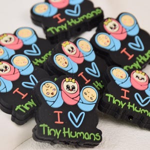May include: A close-up of several black silicone charms with a cartoon image of two babies in swaddles. The charms have a heart and the text "I love Tiny Humans".