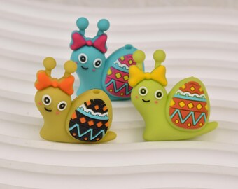 Puede incluir: Tres figuras de caracoles coloridos. Uno es azul claro con un lazo rosa, los otros dos son amarillos y verdes con lazos naranjas y amarillos. Cada caracol tiene una concha estampada con una cara sonriente.