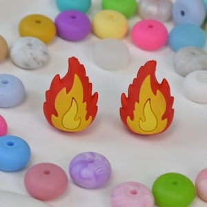 May include: Two red and yellow flame-shaped charms are the focal point, surrounded by a variety of colorful beads in shades of pink, blue, green, and yellow. The beads are round with a hole through the center, suggesting they are for jewelry making.