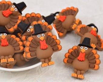 May include: A group of brown turkey-shaped decorations with orange and brown tail feathers, orange feet, and a red wattle. Each turkey wears a black top hat. The turkeys are in a white bowl and scattered on a white surface.