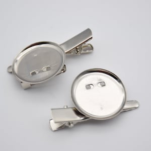 May include: Two silver metal badge clips with round, blank, silver metal bases. The bases have two small holes for attaching a pin or other fastener.