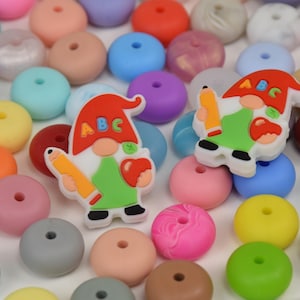 Peut inclure: Un assortiment de perles en silicone colorées dans diverses teintes, dont le rose, le bleu, le vert et l'orange. Deux breloques en forme de gnome avec des crayons et les lettres "ABC" sont également présentes. Ces perles sont adaptées à la création.