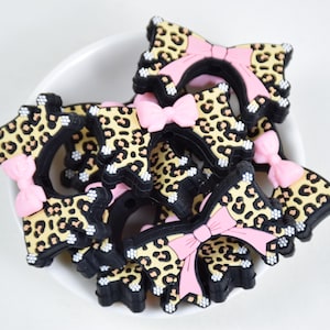 Op de afbeelding: Een verzameling accessoires in de vorm van een luipaardprint met zwarte randen en roze strikken. De accessoires liggen in een witte kom, wat een speelse en modieuze uitstraling creëert. Dit kunnen haaraccessoires of decoratieve versieringen zijn.