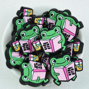 Op de afbeelding: Een verzameling groene, kikker-vormige bedels met zwarte contouren, elk met een roze boek en een drankje. Op het boek staat "DON'T JUDGE ME I'LL EAT YOU". De bedels liggen in een witte kom.