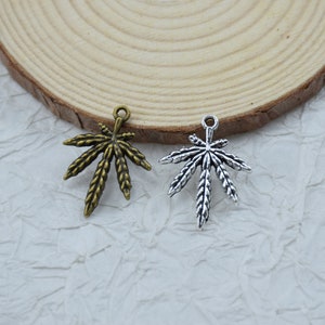 Op de afbeelding: Twee metalen bladbedels, één in antiek goud en de andere in zilver, worden getoond op een wit oppervlak met textuur. De bedels zijn gedetailleerd met aderen en hebben een lus om aan sieraden of andere knutselwerken te bevestigen.