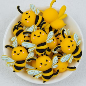 May include: A collection of yellow and black bee-shaped embellishments with smiling faces and light blue wings, arranged in a white bowl. The bees have black stripes and antennae. The image is taken from a high angle.