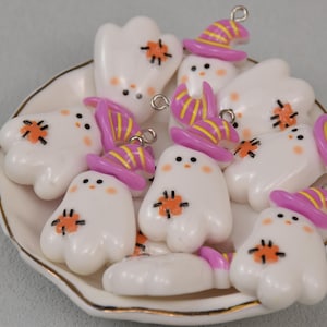 Op de afbeelding: Een verzameling witte spookvormige bedels met oranje en zwarte accenten, elk versierd met een kleine, roze en geel gestreepte heksenhoed. De bedels zijn gerangschikt op een wit bord met een gouden rand, perfect voor Halloween-thema knutselen.