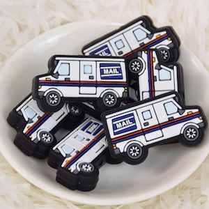 Op de afbeelding: Een groep van zes witte postwagens met blauwe en rode strepen en het woord "MAIL" erop gedrukt. De wagens zijn gemaakt van zwart rubber en zijn bedoeld om te worden gebruikt als schoenbedels.