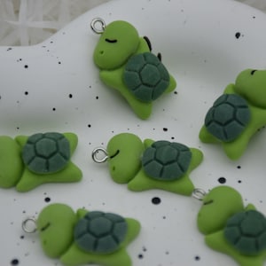 Puede incluir: Una colección de pequeños abalorios de tortuga verde con caparazones texturizados de color verde oscuro. Cada abalorio tiene un lazo plateado en la parte superior para sujetarlo a joyas u otras manualidades. Las tortugas están tumbadas con los ojos cerrados.