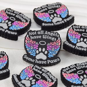 Puede incluir: Un conjunto de seis dijes de goma negra con un diseño de huella de pata arcoíris y el texto "Not all Angels have Wings, Some have Paws".