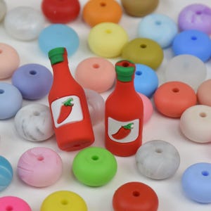 Op de afbeelding: Een verzameling kleurrijke kralen in verschillende tinten roze, blauw, groen, oranje en wit, samen met twee rode flesvormige bedels met een chilipeperlabel. Deze kralen zijn geschikt voor het maken van sieraden en knutselen.