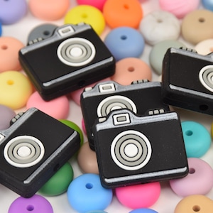 Peut inclure: Gros plan sur cinq perles en silicone noires en forme d'appareil photo avec des détails blancs. Les perles sont éparpillées sur une surface avec des perles en silicone colorées.