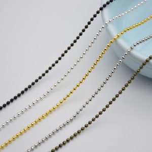 May include: Four strands of ball chain in different colors: black, silver, gold, and bronze. The chains are laid out in a diagonal line on a white surface.