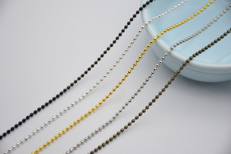 May include: Four strands of ball chain in different colors: black, silver, gold, and bronze. The chains are laid out in a diagonal line on a white surface.