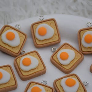Puede incluir: Nueve abalorios cuadrados con un diseño de huevo frito sobre tostadas. Los abalorios están hechos de arcilla polimérica marrón, amarilla y blanca y tienen anillas de salto plateadas.