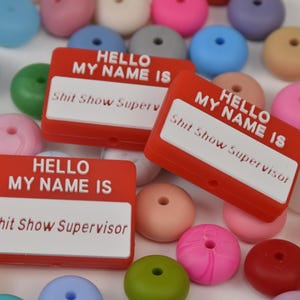May include: Red name tags with the text "HELLO MY NAME IS" and "Shit Show Supervisor" in white. The tags are surrounded by colorful, round beads in various shades of pink, blue, green, and orange.