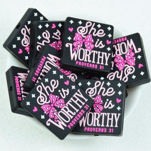 Può includere: Magneti quadrati neri con la scritta "She is WORTHY" in rosa e bianco, con un fiocco rosa e cuori. È incluso anche il testo "PROVERBI 31". I magneti sono ammucchiati in una ciotola bianca.