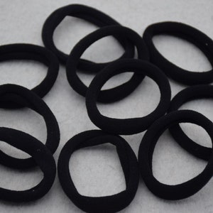 Puede incluir: Un grupo de diez gomas para el pelo negras, hechas de tela suave y elástica. Las gomas para el pelo están dispuestas en un círculo sobre una superficie gris.