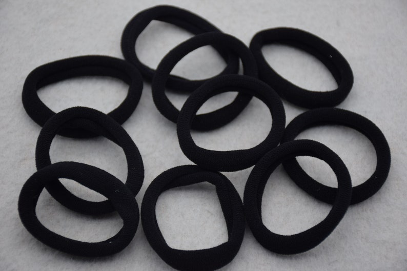 May include: A group of ten black hair ties, made of soft, stretchy fabric. The hair ties are arranged in a circle on a gray surface.