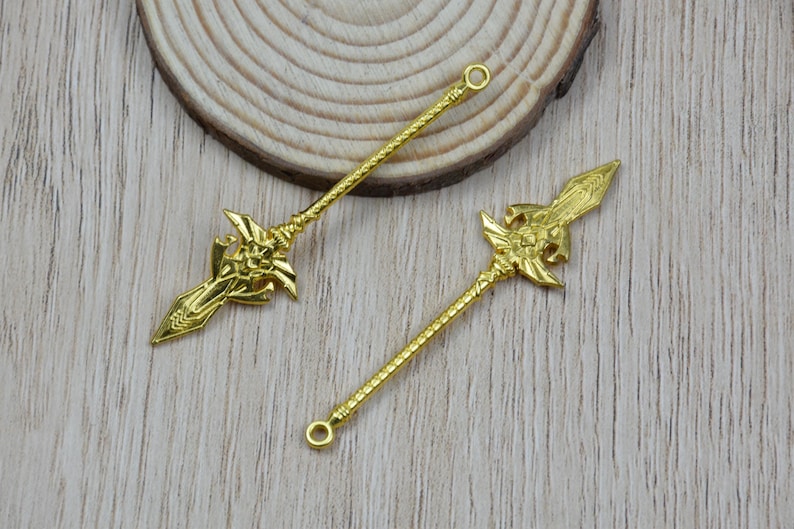 K&ouml;nnte beinhalten: Zwei goldfarbene Speer-Anh&auml;nger mit aufwendigen Details. Jeder Anh&auml;nger hat ein verziertes Speerkopf-Design und einen strukturierten Schaft. Die Anh&auml;nger haben oben eine Schlaufe zur einfachen Befestigung, ideal f&uuml;r Schmuckherstellung oder Bastelprojekte.