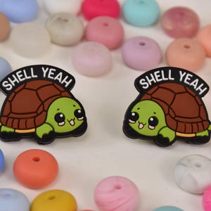 Könnte beinhalten: Zwei schildkrötenförmige Anhänger mit dem Text "SHELL YEAH" darüber. Die Schildkröten haben braune Panzer, grüne Körper und lächelnde Gesichter. Der Hintergrund ist mit bunten, runden Perlen in verschiedenen Pastelltönen gefüllt.