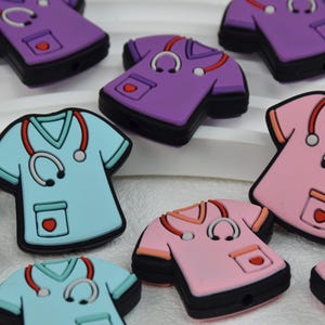 Puede incluir: Surtido de batas de quirófano de silicona de colores vivos con un diseño de estetoscopio. Las batas son en tonos de morado, rosa y azul.