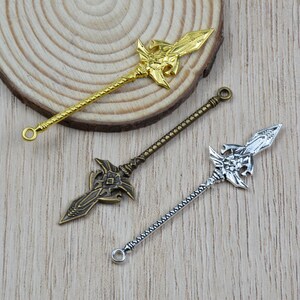 K&ouml;nnte beinhalten: Drei dekorative Speer-Anh&auml;nger in Gold-, Bronze- und Silberfarben. Jeder Anh&auml;nger hat ein verziertes Speerkopf-Design mit einem detaillierten Schaft und einer kleinen Schlaufe am Ende. Die Anh&auml;nger werden auf einer hellen Holzoberfl&auml;che pr&auml;sentiert.