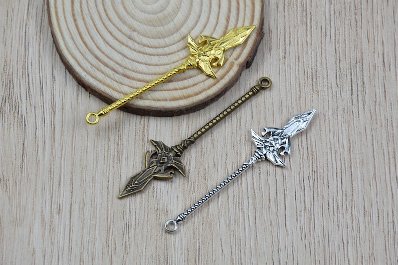 K&ouml;nnte beinhalten: Drei dekorative Speer-Anh&auml;nger in Gold-, Bronze- und Silberfarben. Jeder Anh&auml;nger hat ein verziertes Speerkopf-Design mit einem detaillierten Schaft und einer kleinen Schlaufe am Ende. Die Anh&auml;nger werden auf einer hellen Holzoberfl&auml;che pr&auml;sentiert.