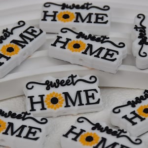 Könnte beinhalten: Acht weiße Silikon-Anhänger mit einer Sonnenblume und dem Text "sweet home" in schwarzer Schrift.