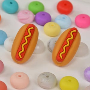 May include: Two orange hot dog-shaped beads with red and yellow details are surrounded by colorful round beads in various colors. The beads are likely for jewelry making or crafts.